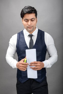 Hintli genç iş adamı resmi bir klasör üzerinde sunum yapıyor. Beyaz gömlek, siyah kravat ve gri arka planda siyah ceket giyiyor. Erkek modeli.