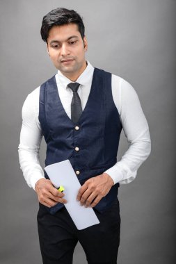 Hintli genç iş adamı resmi bir klasör üzerinde sunum yapıyor. Beyaz gömlek, siyah kravat ve gri arka planda siyah ceket giyiyor. Erkek modeli.