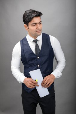 Hintli genç iş adamı resmi bir klasör üzerinde sunum yapıyor. Beyaz gömlek, siyah kravat ve gri arka planda siyah ceket giyiyor. Erkek modeli.