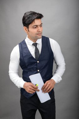 Hintli genç iş adamı resmi bir klasör üzerinde sunum yapıyor. Beyaz gömlek, siyah kravat ve gri arka planda siyah ceket giyiyor. Erkek modeli.