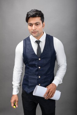 Hintli genç iş adamı resmi bir klasör üzerinde sunum yapıyor. Beyaz gömlek, siyah kravat ve gri arka planda siyah ceket giyiyor. Erkek modeli.