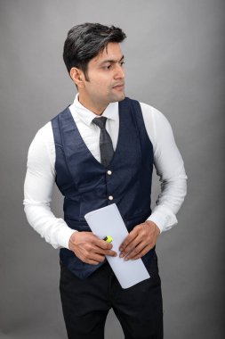 Hintli genç iş adamı resmi bir klasör üzerinde sunum yapıyor. Beyaz gömlek, siyah kravat ve gri arka planda siyah ceket giyiyor. Erkek modeli.