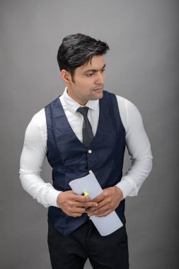 Hintli genç iş adamı resmi bir klasör üzerinde sunum yapıyor. Beyaz gömlek, siyah kravat ve gri arka planda siyah ceket giyiyor. Erkek modeli.