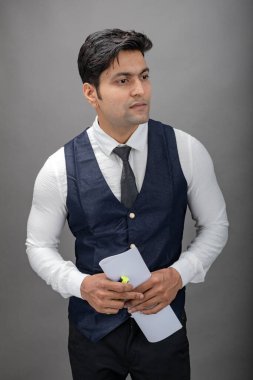 Hintli genç iş adamı resmi bir klasör üzerinde sunum yapıyor. Beyaz gömlek, siyah kravat ve gri arka planda siyah ceket giyiyor. Erkek modeli.