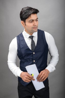 Hintli genç iş adamı resmi bir klasör üzerinde sunum yapıyor. Beyaz gömlek, siyah kravat ve gri arka planda siyah ceket giyiyor. Erkek modeli.