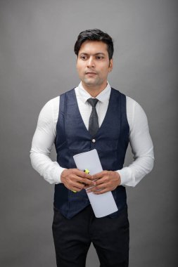 Hintli genç iş adamı resmi bir klasör üzerinde sunum yapıyor. Beyaz gömlek, siyah kravat ve gri arka planda siyah ceket giyiyor. Erkek modeli.
