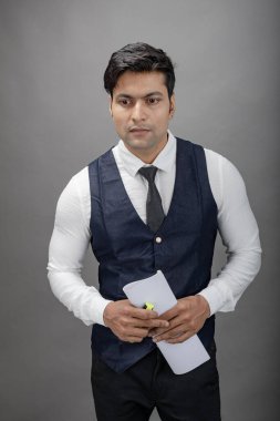 Hintli genç iş adamı resmi bir klasör üzerinde sunum yapıyor. Beyaz gömlek, siyah kravat ve gri arka planda siyah ceket giyiyor. Erkek modeli.