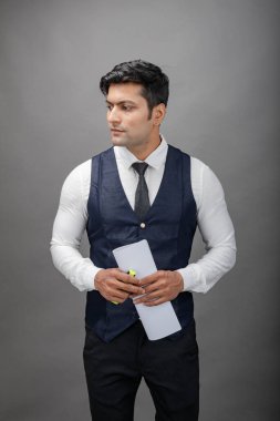 Hintli genç iş adamı resmi bir klasör üzerinde sunum yapıyor. Beyaz gömlek, siyah kravat ve gri arka planda siyah ceket giyiyor. Erkek modeli.