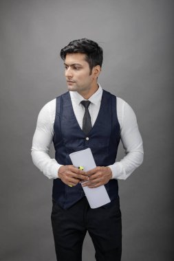 Hintli genç iş adamı resmi bir klasör üzerinde sunum yapıyor. Beyaz gömlek, siyah kravat ve gri arka planda siyah ceket giyiyor. Erkek modeli.