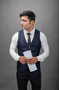 Hintli genç iş adamı resmi bir klasör üzerinde sunum yapıyor. Beyaz gömlek, siyah kravat ve gri arka planda siyah ceket giyiyor. Erkek modeli.