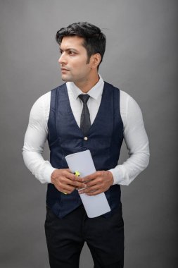 Hintli genç iş adamı resmi bir klasör üzerinde sunum yapıyor. Beyaz gömlek, siyah kravat ve gri arka planda siyah ceket giyiyor. Erkek modeli.