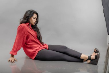 Gri arka planda poz veren gündelik giyimli Hintli genç bayan manken. Kırmızı gömlek, siyah kot pantolon ve siyah topuklu uzun siyah saçlı bir manken..