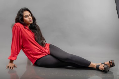 Gri arka planda poz veren gündelik giyimli Hintli genç bayan manken. Kırmızı gömlek, siyah kot pantolon ve siyah topuklu uzun siyah saçlı bir manken..