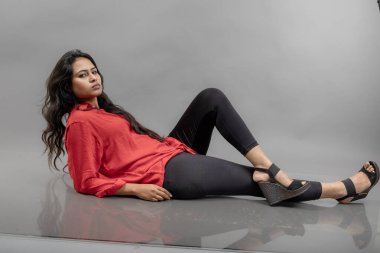 Gri arka planda poz veren gündelik giyimli Hintli genç bayan manken. Kırmızı gömlek, siyah kot pantolon ve siyah topuklu uzun siyah saçlı bir manken..