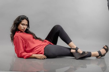 Gri arka planda poz veren gündelik giyimli Hintli genç bayan manken. Kırmızı gömlek, siyah kot pantolon ve siyah topuklu uzun siyah saçlı bir manken..