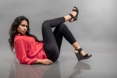 Gri arka planda poz veren gündelik giyimli Hintli genç bayan manken. Kırmızı gömlek, siyah kot pantolon ve siyah topuklu uzun siyah saçlı bir manken..