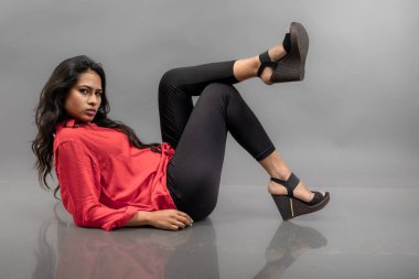 Gri arka planda poz veren gündelik giyimli Hintli genç bayan manken. Kırmızı gömlek, siyah kot pantolon ve siyah topuklu uzun siyah saçlı bir manken..