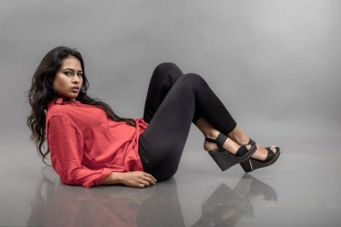 Gri arka planda poz veren gündelik giyimli Hintli genç bayan manken. Kırmızı gömlek, siyah kot pantolon ve siyah topuklu uzun siyah saçlı bir manken..