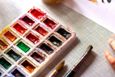 Paletlere odaklı suluboya bir kutu, iki fırça, gün ışığıyla gri bir masanın üzerine çizen bir çocuk parçası..