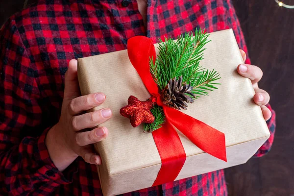 Kerst nieuwjaar cadeau, verpakt in ambachtelijk gewoon papier en versierd met rood satijnen lint met sparren takken en een speelgoed in de handen van een meisje kind op een donkere achtergrond. — Stockfoto