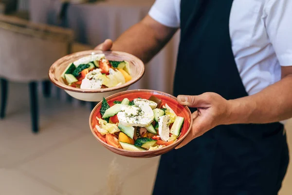 Ensalada griega con queso feta, tomates jugosos, pimiento rojo, pepino y lechuga. El camarero sostiene los platos en sus manos.. —  Fotos de Stock