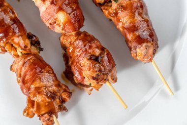 Izgara domuz pastırmasına sarılmış tavuk eti beyaz bir tabakta sunuluyor. Tahta şiş-2 'ye asılmış..