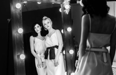 Seksi genç sarışın ve esmer aynaya bakıyor. Deri mücevherler ve kocaman altın küpeler..