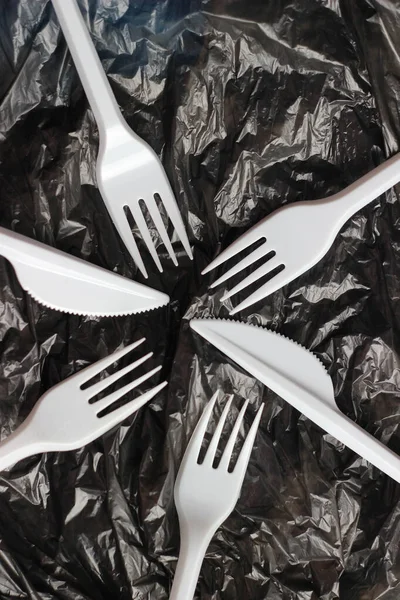 Plastic utensils, white forks and knives on a black plastic bag. Abstract background. Environmental pollution, environmental problems