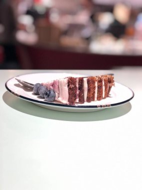 Beyaz bir masada çay kaşığı ile mavi böğürtlenli bir parça sünger kek beyaz bir tabakta yatıyor.