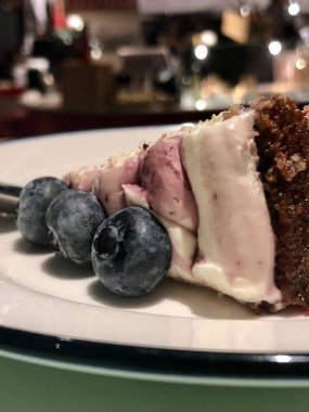 Lokantada çay kaşığı ile beyaz bir tabakta üç mavi böğürtlenli pandispanya dilimi.