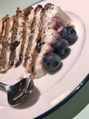 Beyaz tabakta üç büyük mavi böğürtlenli bir dilim kek, sol alt köşede bir çay kaşığı, yakın plan.