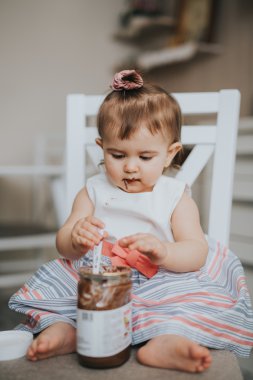 Sevimli küçük bebek kız mutfakta masada kaşık çikolata krema ile yemek yiyen ve komik mutlu yüzler yapar 