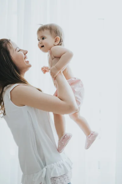 Beautiful mother portrait playing with her cute little girl in a white room in the morning — Stock Photo, Image