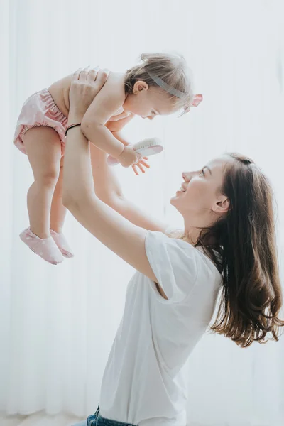 Beautiful mother portrait playing with her cute little girl in a white room in the morning — Stock Photo, Image