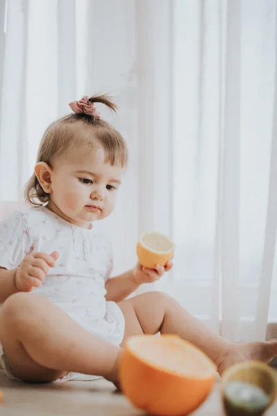 Cute little baby girl on the table in the kitchen eats citrus fruits full of vitamins and makes funny cute faces — Stock Photo, Image