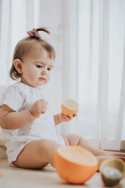 Милая маленькая девочка на столе на кухне ест цитрусовые, полные витаминов и делает смешные милые лица — стоковое фото