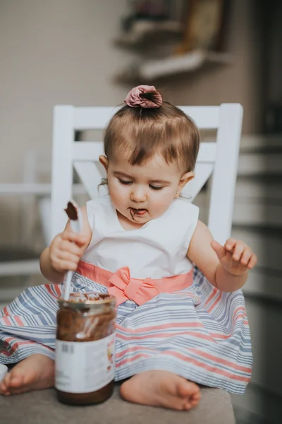 Милая маленькая девочка на столе на кухне ест с ложкой шоколадного крема и делает смешные счастливые лица — стоковое фото