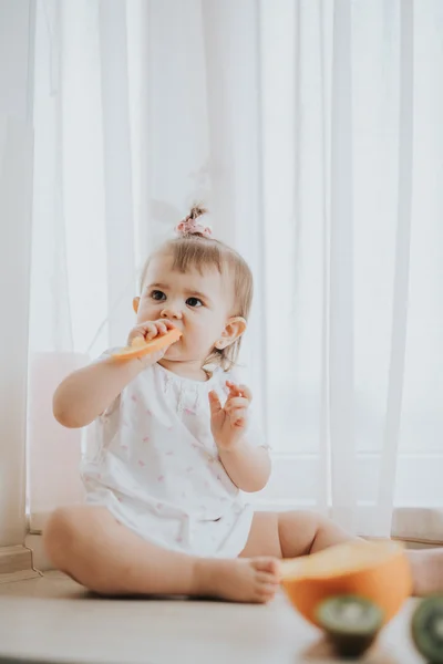 Cute little baby girl on the table in the kitchen eats citrus fruits full of vitamins and makes funny cute faces — Stock Photo, Image