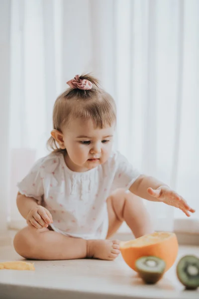 Linda niña en la mesa en la cocina come cítricos llenos de vitaminas y hace caras lindas divertidas —  Fotos de Stock