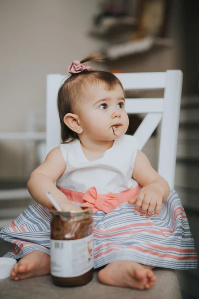 Cute little baby girl on the table in the kitchen eats with the spoon chocolate cream and makes funny happy faces