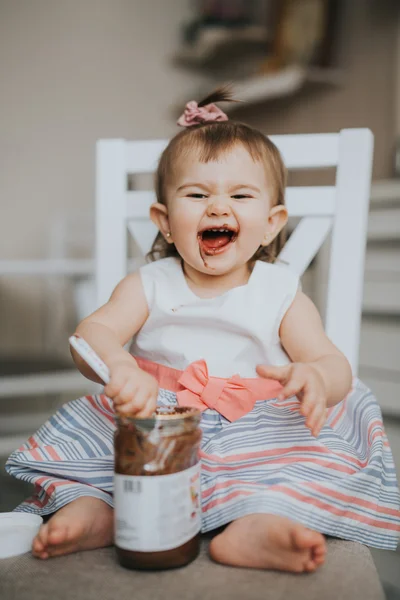 Милая маленькая девочка на столе на кухне ест с ложкой шоколадного крема и делает смешные счастливые лица — стоковое фото