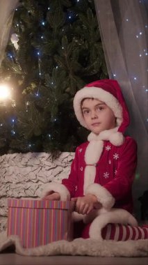 Noel Baba kostümlü çocuk, süpriz bir şekilde süslü bir Noel ağacının yanında ışıldayan bir hediye kutusu açıyor. Yüksek kalite 4k görüntü