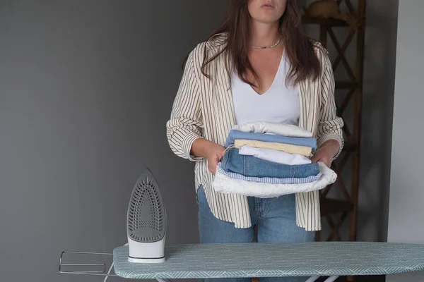 Kadın, elinde ütüyle evde çamaşır ütülüyor ve günlük ev işleri konseptini gösteren giysileri düzenli bir şekilde istifliyor.
