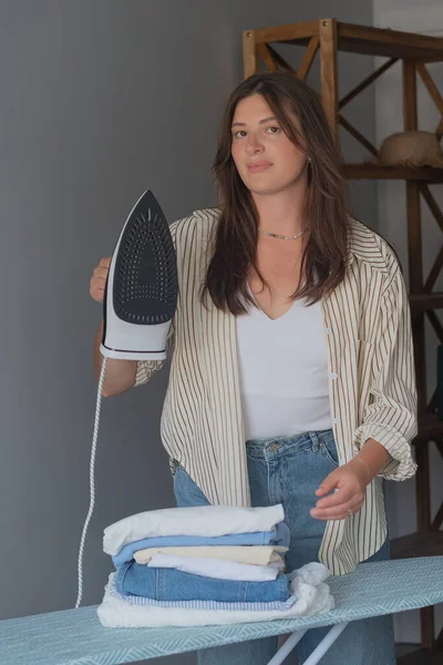 Gömlekli ve kot pantolonlu bir kadın buhar ütüsüyle giysilerini ütülüyor ve sabah rutini ve düzeni konseptine hazırlanıyor.