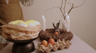 Önlüklü ve başörtülü bir kadın Paskalya masasına eğilip yumurta ve pastanın yanındaki çelenge mum üflüyor. Yüksek kalite 4k görüntü