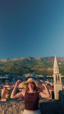 Hasır şapkalı ve etekli bir kız Budva Kalesi 'nin tarihi çatıları ve Vaftizci Aziz John kilisesi olan eski şehrin panoramik manzarasının tadını çıkarıyor. Yüksek kalite 4k görüntü