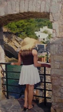 Hasır şapkalı ve taş kemerli, pofuduk etekli kadın kameralı zoom, turkuaz deniz kayalıkları ve küçük Petrovac kalesi Karadağ 'ı gözler önüne seriyor. Yüksek kalite 4k görüntü