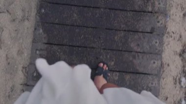 Yazlık sandaletli bir kızın güneşli sahil ve kıyı şeridi boyunca ahşap patikada yürüdüğü ilk kişi. Yüksek kalite 4k görüntü
