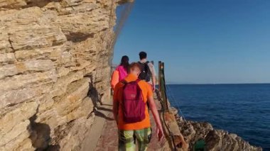 Budva, Karadağ 10 Eylül 2025, Sırt çantalı turistlerin parlak yaz kıyafetleri içinde deniz kıyısında yürüyüş yaparken manzaranın tadını çıkarmaları. Yüksek kalite 4k görüntü
