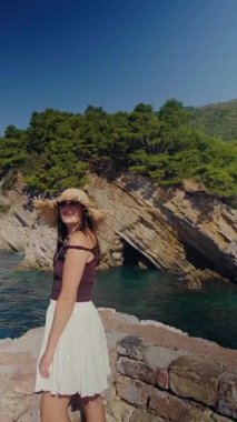 Hasır şapkalı ve beyaz etekli bir kadın yüksek uçurumların manzarasını izliyor yeşil ağaçlar ve Adriyatik dalgaları özgürlük hissi veriyor. Yüksek kalite 4k görüntü